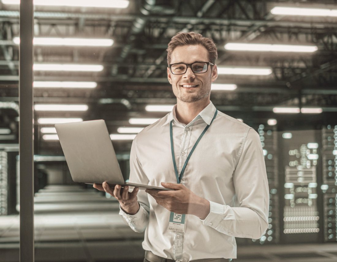 A smiling IT engineer holding a laptop in a server room, network and storage racks glowing behind him