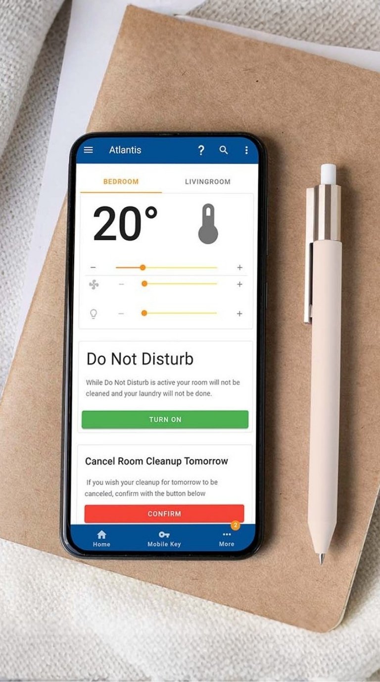 A smartphone resting on a notebook beside a coffee cup, showing the hotel app's in-room control screen with temperature, Do Not Disturb and a Mobile Key tab