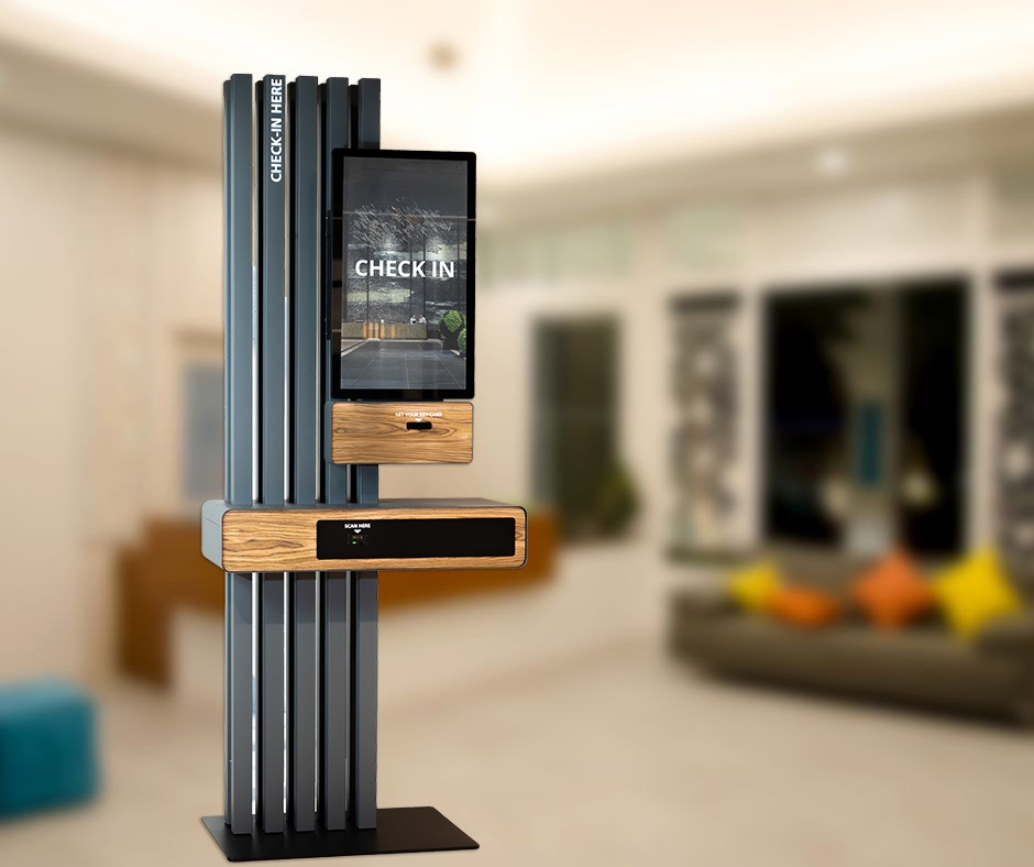 A branded self-check-in kiosk standing in a bright hotel lobby, with a CHECK IN screen and key-card dispenser