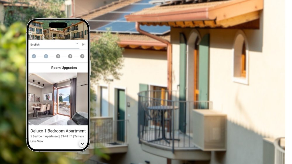 A smartphone showing a pre-arrival room-upgrade step — a Deluxe 1 Bedroom Apartment with lake-view terrace — against a sunlit Lake Garda hotel facade