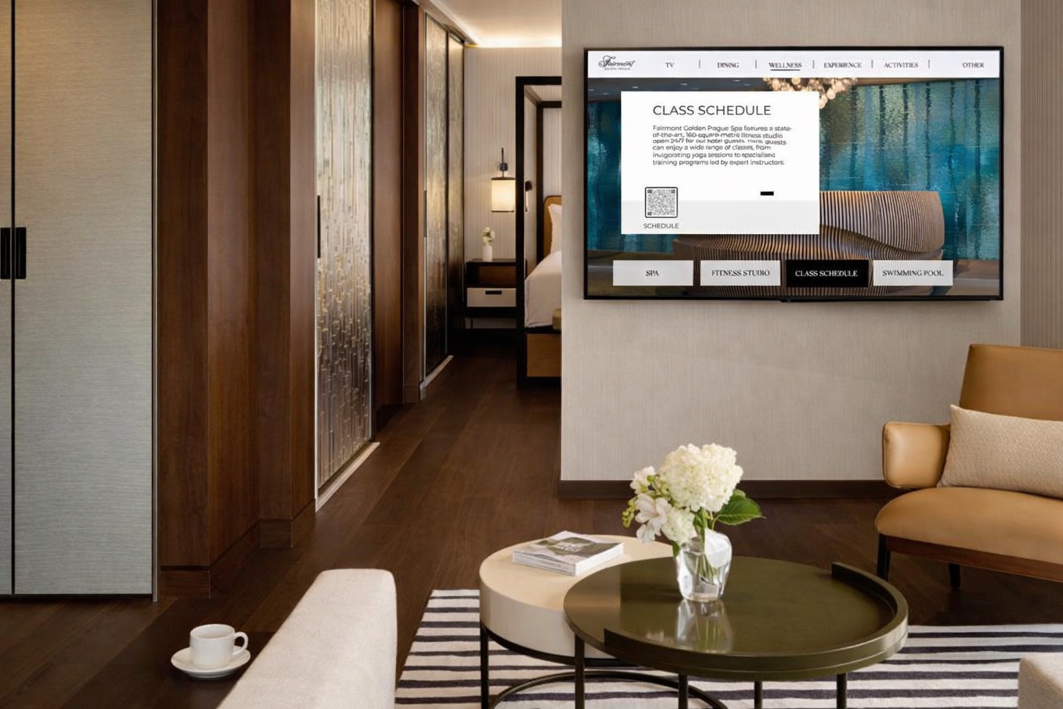 A guest-room wall-mounted TV showing the in-room wellness menu with a class schedule and QR code, in a warm suite with a hallway view to the bedroom